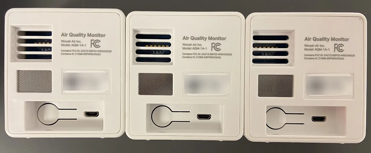 Back view of three Woosh Air Quality Monitor units, model AQM-1A-1, showing the FCC marking, sensor vents and mounting slot; serial and pairing labels are blurred