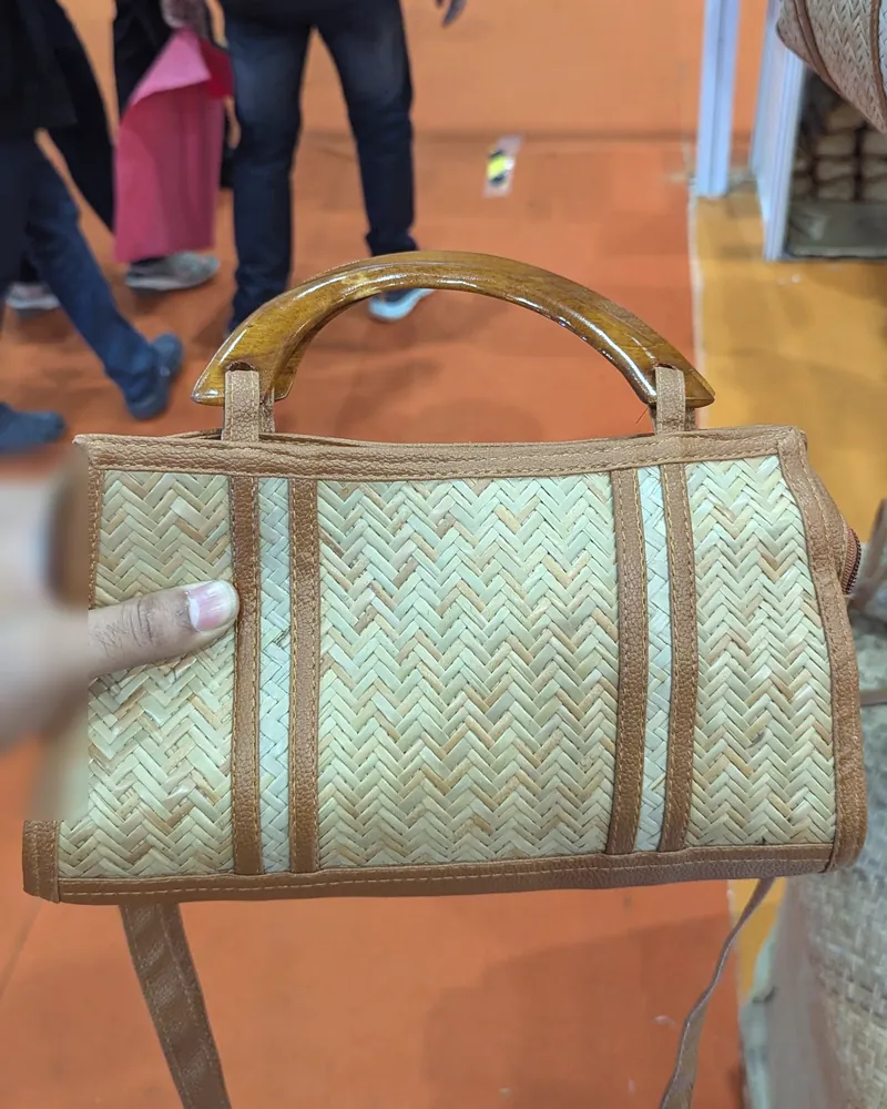 A hand holding a woven cane handbag with tan leather trim and a wooden handle.