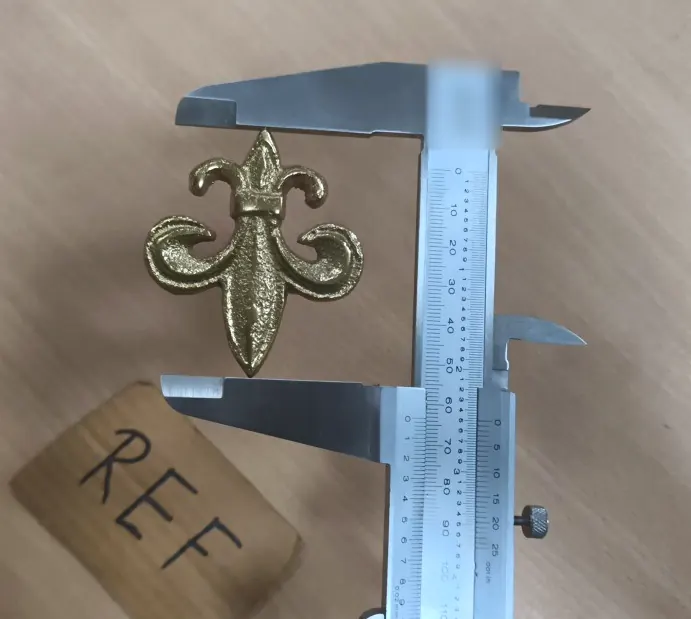 A cast brass fleur-de-lis motif measured between the jaws of a vernier caliper.