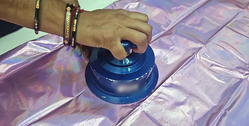 A hand presses a round GSM sample cutter onto a sheet of iridescent pink fabric.