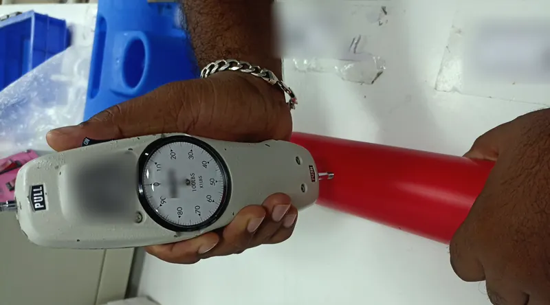 An inspector presses a dial push-pull force gauge against the end of a red moulded plastic handle.