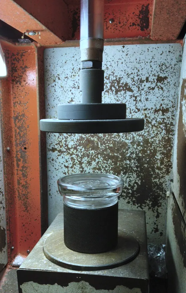 A clear glass jar on a knurled stand under the steel platen of a vertical load tester.