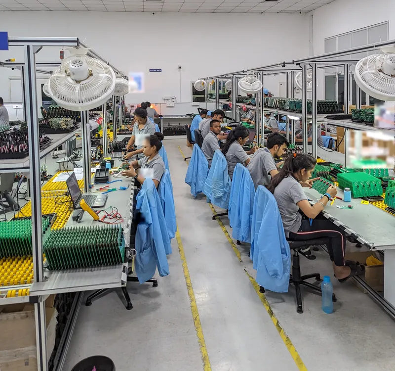 Two long rows of assembly benches with operators in grey polos working on circuit boards, blue ESD smocks draped over their chairs.