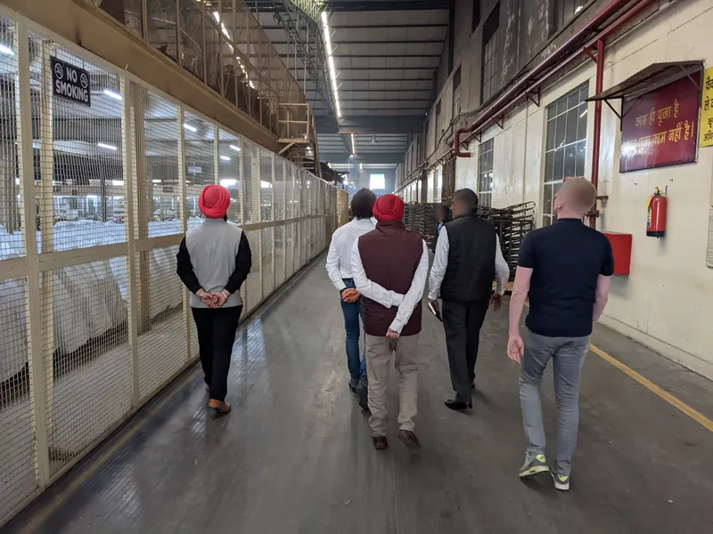 Five people seen from behind walking down a long factory aisle beside a mesh-fenced store; faces blurred.